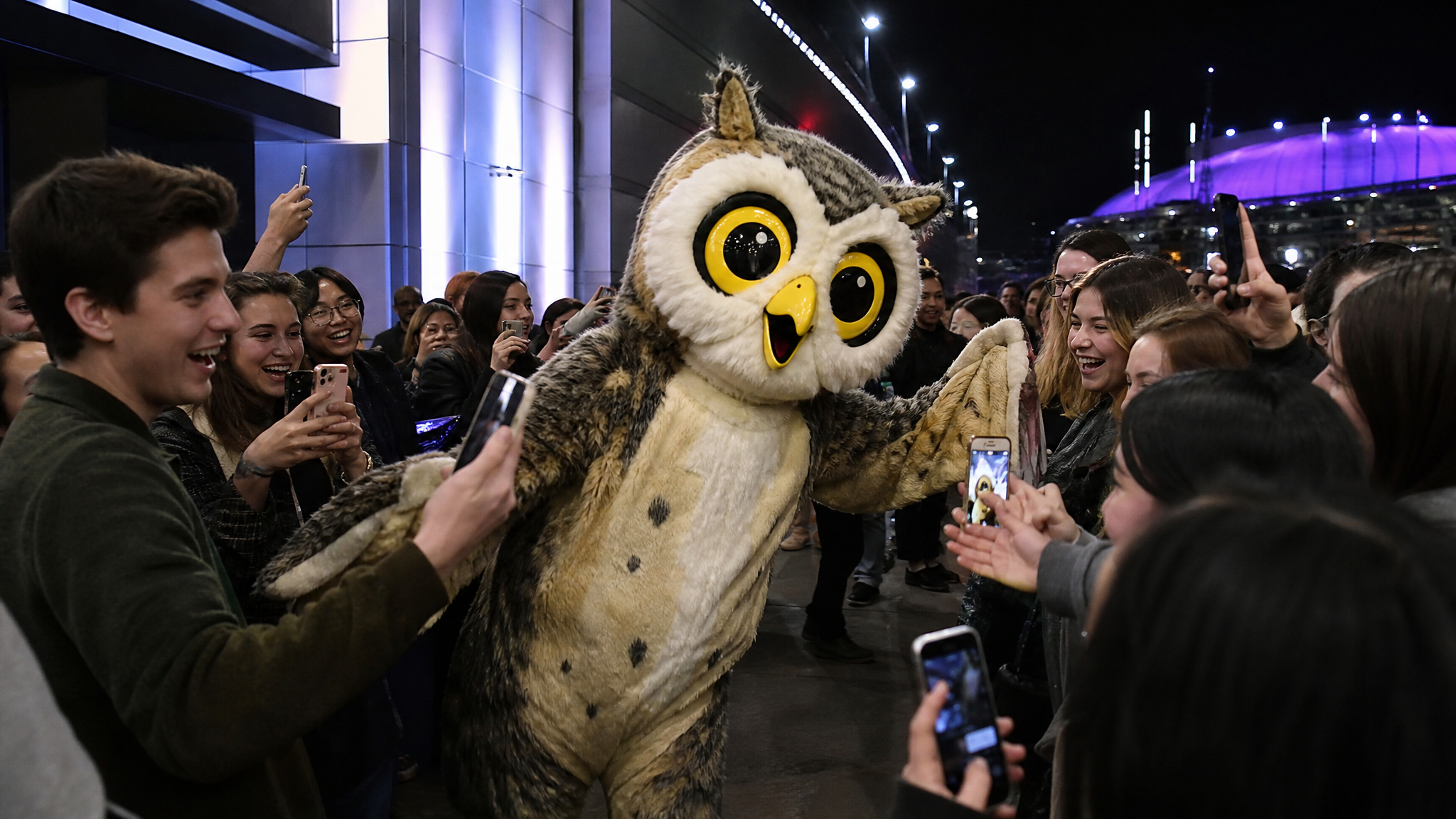 A person in a bright owl-like mascot costume moving through a crowd outside an entertainment venue while people film on their phones